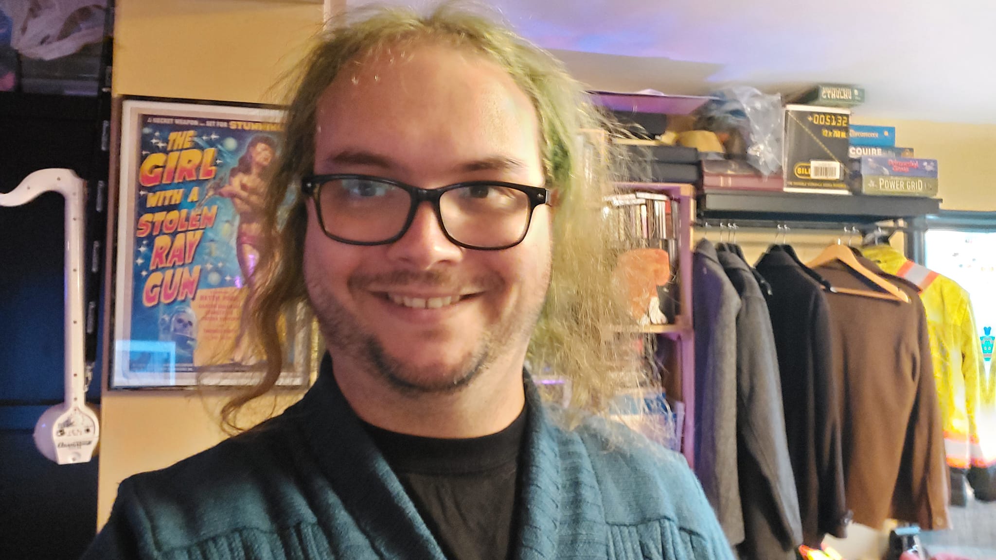 A man with long green hair wears a black shirt and dark green cardigan, behind him is a poster for a fake movie called "Girl with the Stolen Raygun"